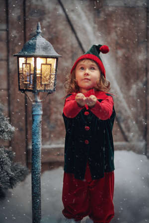 Winter Christmas tale. A little cute elf girl catches snowflakes and rejoices, standing at a wooden hut and a snow-covered lantern under a snowfall.の写真素材