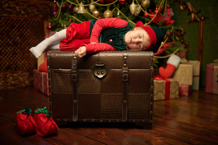 Christmas tale. A little elf girl looks out of a large chest in a vintage room fabulously decorated for Christmas. Beautiful Christmas tree in the background.の写真素材