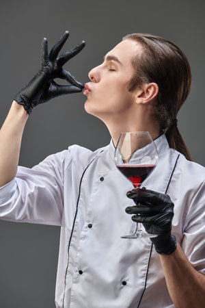 Professional sophisticated male chef in neat uniform examines excellent red wine in a glass. Studio shot on a grey background.の写真素材