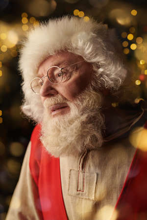 Christmas traditions and fairy tales. A portrait of a kind Santa Claus in a canvas shirt, a red vest and a hat in his cozy home decorated with shining Christmas lights.の写真素材