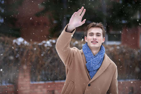 Joyful handsome young man in a trendy coat and scarf stands on the city street by a brick house waving his hand to the camera. Men's fashion and lifestyle.の写真素材