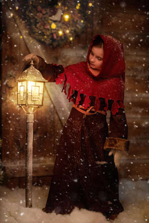 Full length shot of a cute little girl in medieval clothes standing by a wooden house and curiously looking at a beautiful burning lantern on a snowy night. Once upon a Christmas.の写真素材