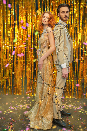 Romantic Christmas and New Year celebration. Beautiful young couple in love in luxury evening dresses stands on the background of shiny gold foil curtains tied with Christmas lights. Holiday fashion.の写真素材