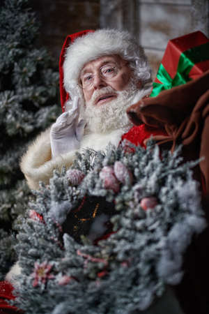 Magical Christmas and New Year. Fabulous Santa Claus stands outside a house holding a heavy bag of presents and a Christmas wreath.の写真素材