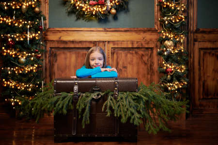 Sweet little girl in a pretty medieval dress joyfully sits in a fabulous Christmas setting of a vintage room leaning on a chest with spruce branches. Christmas dreams and wishes.の写真素材