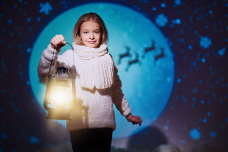 Lovely blonde girl in a warm sweater and scarf stands on the background of the moon and Santa Claus flying across the sky with a lantern. Winter kid's fashion. Merry Christmas and Happy New Year!の写真素材