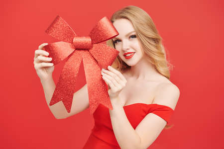 Pretty blonde girl with bright evening make-up in elegant red dress poses with a festive red bow on a red background. Fashion and beauty. Make-up and cosmetics. Holiday celebration.の写真素材