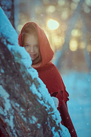 A cheerful medieval girl in caftan and cape joyfully peeks out from behind a tree in a snowy forest at the sunset. Fairy tales on Christmas and New Year.の写真素材