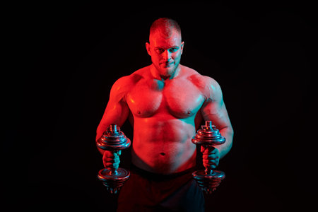 Strong, athletic man training hard with dumbbells on a black background with red light. Male beauty...
