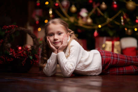 A happy little girl in a white winter sweater and red pants lying on the floor and dreamily looking at the camera. Festive Christmas interior background. New Year and Christmas dreams.の写真素材