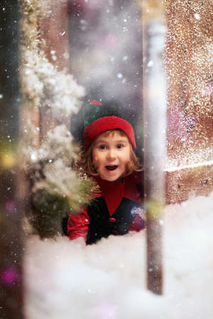 Time of Christmas miracles. Portrait of a cute little elf girl looking out excitedly from behind the door of a snow-covered wooden house in a fabulous winter forest.の写真素材