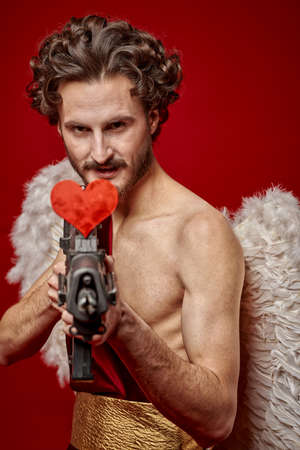 Modern Cupid with a big gun resolutely looking for a victim of love. Red background. God of Love. Valentine's Day and Love.の写真素材