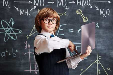 Portrait of an excited clever little boy in school uniform and glasses pressing keys on his laptop in the background of a blackboard with scientific formulas. Smart children. Education.の写真素材