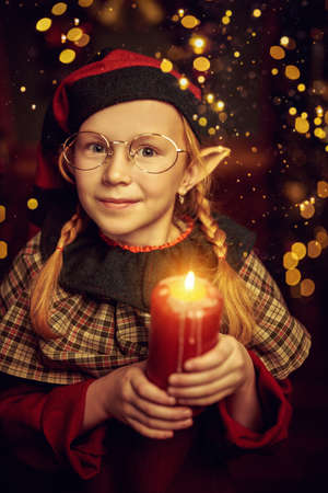 Christmas miracles and dreams. A cute joyful girl elf stands in a night cozy room with shining magical Christmas lights around.の写真素材