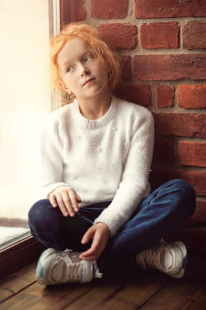 A portrait of a cute red-haired little girl in casual clothes sitting on the windowsill and looking thoughtfully out the window. Natural lighting. Children' fashion. Childhood and emotions.の写真素材