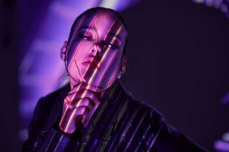 Attractive confident brunette girl in a black leather jacket posing in a dark space with neon beams of light on her face. Virtual reality concept. Futuristic style and beauty, fashion.の写真素材