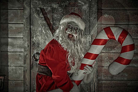 Mad zombie Santa Claus jumps on a Christmas candy cane in a night winter forest near a wooden hut and laughs uncontrollably and evilly. Nightmare at Christmas. Halloween.の写真素材