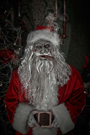 Horror Christmas and New Year.  Scary old man zombie Santa Claus smokes a cigar and smiles evilly while standing in a dark Christmas room. Halloween.の写真素材