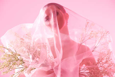 A beautiful graceful girl with small white flowers in her hands, covered with transparent organza in a delicate pink light on a pink background. Cosmetics and perfumery. Skin and body care.の写真素材