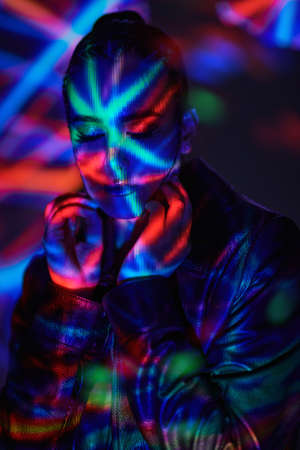 Virtual reality concept. Portrait of an attractive brunette girl with bright makeup and closed eyes in a black leather jacket standing in a dark space filled with coloured lazer beams.の写真素材