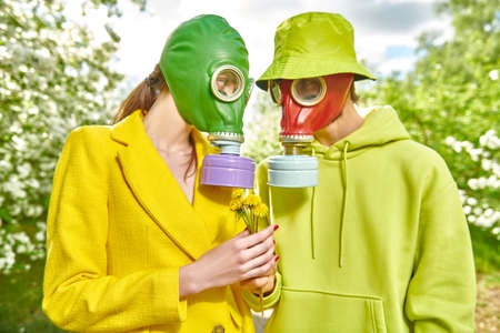 A loving man gives a young woman wild flowers in the park on a sunny day. Couple in love dressed in bright clothes and gas masks. Air pollution, environmental disaster.の写真素材
