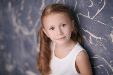Adorable little girl in a white top stands against a grunge wall background smiling slightly. Children's emotions.の写真素材