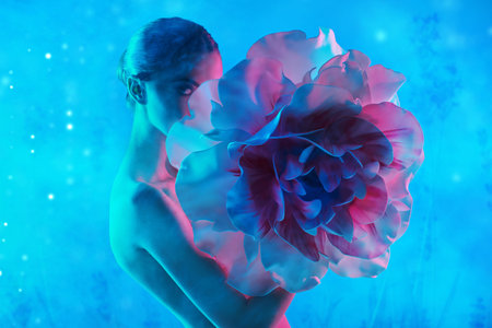 A beautiful naked young woman  poses with a large delicate peony flower, standing in a light haze on a blue background. Beauty portrait. Makeup and cosmetics. Skin and body care.の写真素材