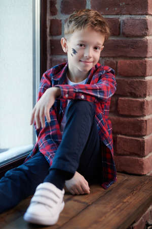 Happy childhood. A stylish carefree boy in a plaid shirt and jeans sits on a windowsill leaning against a brick wall smiling cutely.の写真素材