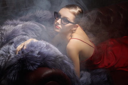 Beautiful glamorous girl in elegant black sunglasses and red satin dress lying in smoke on a leather...