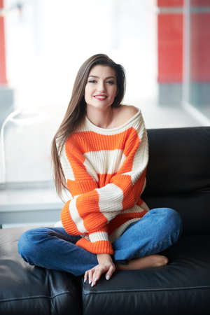 Portrait of a beautiful smiling young woman sitting on a leather sofa in her cozy living room. Happy life, happy people concept.の写真素材