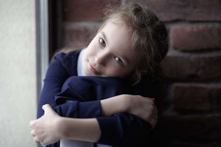 A portrait of a cute girl in school uniform who sits on the windowsill against a brick wall hugging her knees and smiling slightly. Natural lighting. Children, childhood, emotions.の写真素材