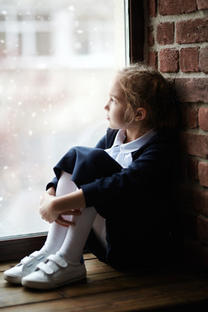 Children, childhood, emotions. A portrait of a girl in school uniform who sits on the windowsill and looks thoughtfully out the window. Natural lighting. Education.の写真素材