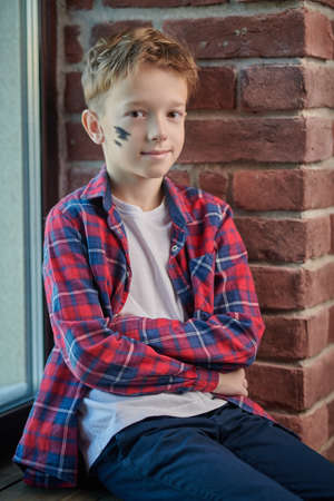 Children's fashion. Portrait of a cute joyful boy in stylish casual clothes sitting relaxed on a windowsill with his arms folded against a brick wall.の写真素材