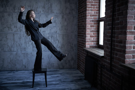 Psychological art portrait. A girl in a black shirt and trousers stands on a stool in a dark room and takes a step towards the window. The human inner world, psychological problems.の写真素材