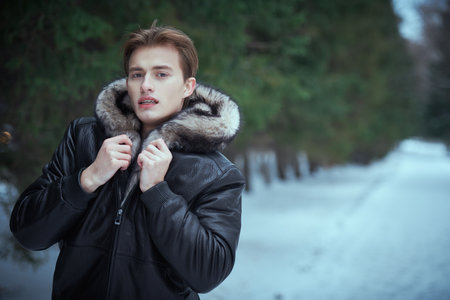 Attractive young blond man in stylish black leather jacket with a fur hood stands in a winter park covered with snow. Men's winter fashion.の写真素材