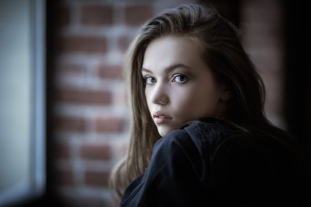 Portrait of a beautiful girl with long hair and in a black shirt, who stands by the window in a room...