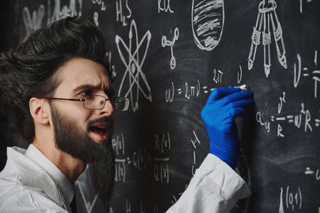 A smart enthusiastic scientist stands at the blackboard explaining  scientific formulas and diagrams. Science and education.の写真素材