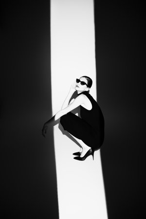 Elegant women's fashion. Portrtait of an elegant girl fashion model posing in a tight black dress and elegant sunglasses in the studio lit by the beam of light. Background with light and shadows.の写真素材