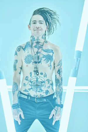 Portrait of a modern expressive punk rock musician with cool mohawk posing with bare torso covered with numerous tattoos. Youth alternative culture. Studio shot on a blue background among neon lamps.の写真素材