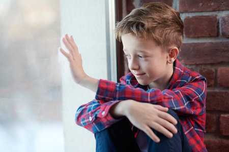 A blond boy in a plaid shirt and jeans sits on a windowsill looking out the window with a sad smile...