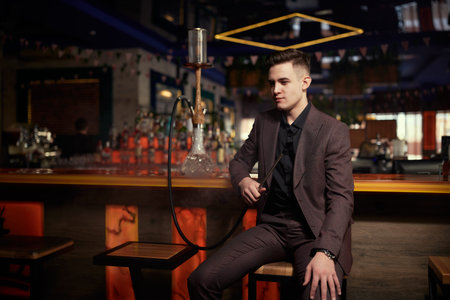 A  handsome well-groomed young man in an expensive suit smoking hookah at the bar in a restaurant....