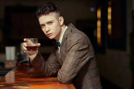 A good-looking young brunette man in a stylish suit and bow tie relaxes sitting at the bar in a...