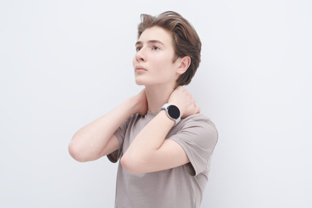 A handsome teenage boy in a grey T-shirt wearily holds his neck with hands and looks pensively away....