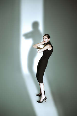 A stunning, elegant female fashion model in a tight black dress and high heels poses in the beam of light. Fashion shot. Light and shadows.の写真素材