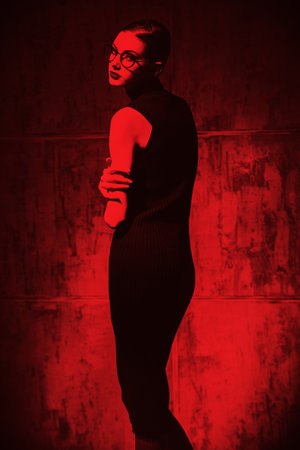Portrait of a pretty elegant girl with bright makeup and stylish glasses posing in black knitted bodycon dress on a grunge background. Red lighting. Fashion shot.の写真素材