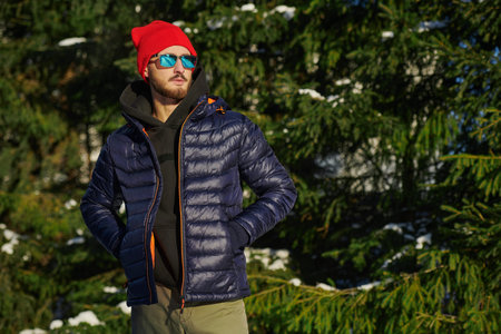 A handsome confident brunette man in a winter down jacket, a knitted hat and sunglasses standing by...