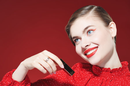 Close up shot of a beautiful elegant woman with bright red arrows and red lipstick holding a tube of red lipstick and smiling charmingly. Burgundy background. Make up and cosmetics.の写真素材