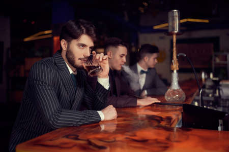 A well-groomed handsome brunette man in an expensive suit rests at the bar in a restaurant drinking whiskey. Businessman. Luxury lifestyle.の写真素材