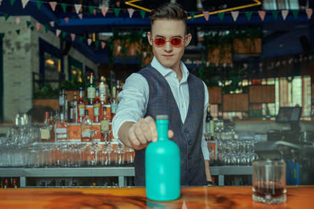 A stylish handsome barman in elegant white shirt and vest serves a drink in a cozy bar.の写真素材