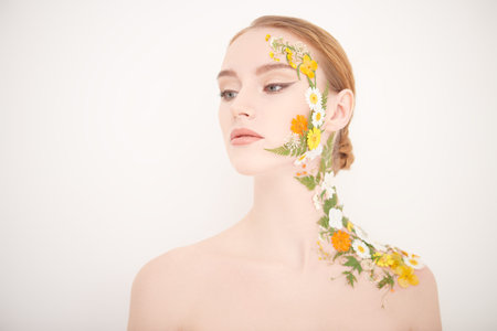 Beauty shot of an attractive red-haired woman with elegant makeup and summer flowers on her face and...